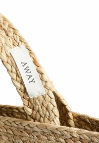 Κοντινή λήψη της λαβής ενός πλεκτού ψάθινου τσάντας με μια λευκή υφασμάτινη ετικέτα ραμμένη πάνω της, που δείχνει τη λέξη "AWAY" με μαύρα γράμματα.