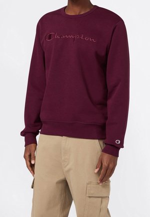 Person trägt einen dunkelroten Champion-Sweatshirt mit gesticktem Logo auf der Brust und beigen Cargohosen, steht vor einem einfarbigen Hintergrund.