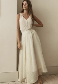 Haut sans manches crème avec des boutons, associé à une longue jupe fluide et texturée de couleur crème. La jupe possède une taille élastique et un design plissé.