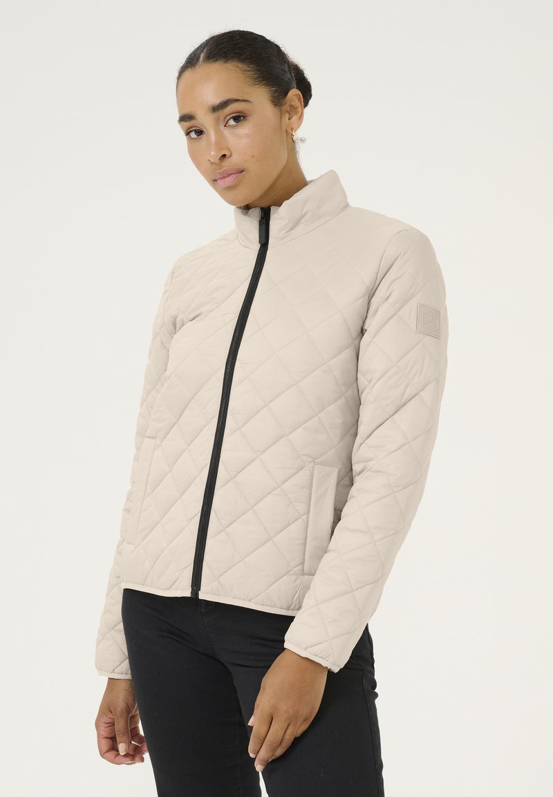 Veste matelassée beige clair avec un col montant, fermeture éclair noire et poches latérales. Présente un motif en losanges et un logo sur la manche gauche.