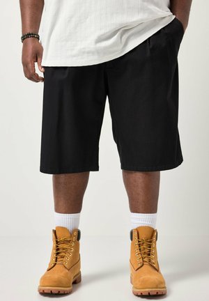 Persona con pantaloncini neri, calzini bianchi, stivali da lavoro color cuoio, una camicia bianca e un braccialetto di perline, in piedi con una mano in tasca.