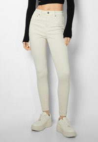 Mujer vistiendo jeans skinny beige de tiro alto, top negro corto de manga larga, y zapatillas de plataforma beige, de pie contra un fondo liso.