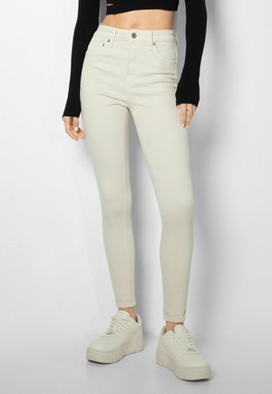 Femme portant un jean skinny beige taille haute, un top noir à manches longues raccourci et des baskets à plateforme beiges, se tenant contre un fond uni.