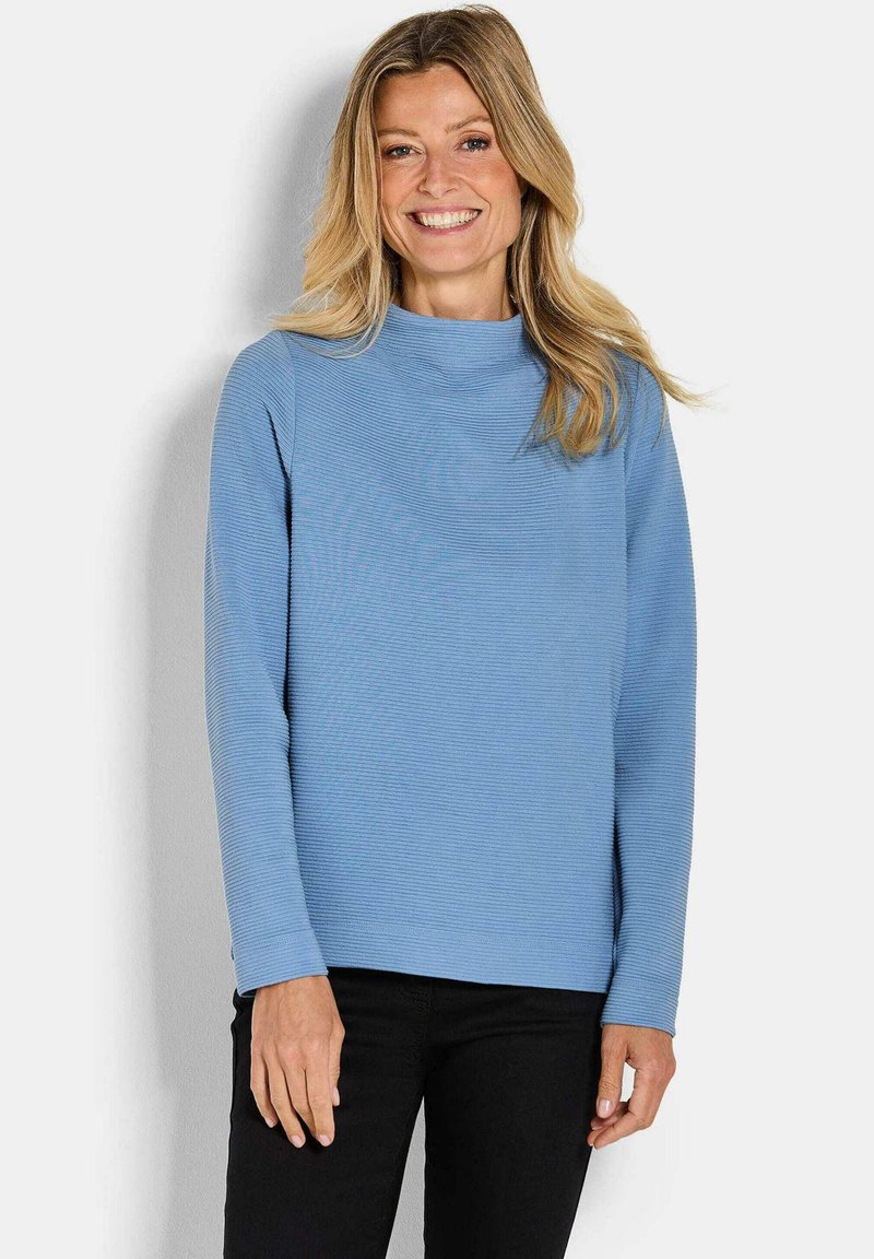Maglia blu a maniche lunghe con collo alto, caratterizzata da una vestibilità relaxed. Indossata con pantaloni neri, il tessuto appare morbido e texture.