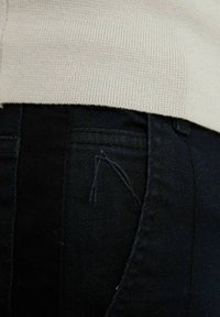 Mustat farkut ja vaaleanbeige neule vyötäröllä. Huomionarvoisia yksityiskohtia ovat näkyvä sauma ja selkeä ompelukuvio taskussa.