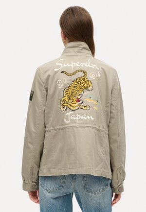 Person iført en beige jakke med broderet tiger og ordene "Superdry Japan" på ryggen, kombineret med blå jeans.