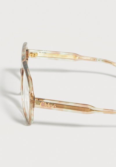 Transparenter beige-goldener Brillenrahmen mit abgerundeten Gläsern und "Chloé"-Logo am Bügel vor weißem Hintergrund.