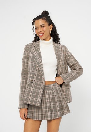 Femme souriante aux cheveux bouclés porte un blazer beige à carreaux et une mini-jupe plissée assortie par-dessus un pull court à col roulé blanc.