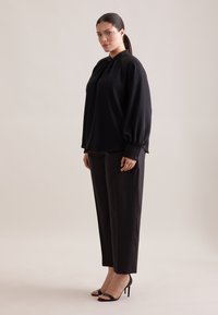 Blusa nera a maniche lunghe con scollatura arricciata, abbinata a pantaloni neri su misura. Presenta polsini con bottone e décolleté neri.