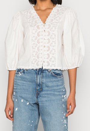 Blouse blanche avec devant en dentelle, boutons en perles et manches bouffantes, associée à un jean taille haute bleu clair avec des éclaboussures de peinture blanche.