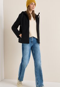 Veste en polaire noire avec capuche, pull crème, jeans en denim bleu et baskets beiges. Modèle portant un bonnet en laine jaune.