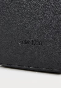 Fekete struktúrált bőrtáska, debosszált „Calvin Klein” logóval, sima szélekkel és minimalista dizájnnal.