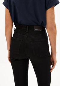 Jeans negros de cintura alta con un acabado texturizado, que cuentan con dos bolsillos traseros y una etiqueta en la cintura. Diseño ajustado en general.