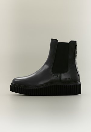 Bottine en cuir noir avec semelle épaisse rainurée, panneau latéral élastique et embossage subtil de la marque près du talon sur un fond neutre.