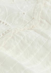 Vitt bomullstyg med texturerad broderi, volangkanter och fin söm med ett subtilt mönster. Mjukt och lätt material.