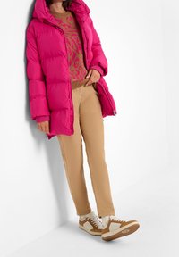 Doudoune rose vif à capuche, manches longues et poignets élastiques ; assortie à un pull beige, un pantalon beige clair et des baskets blanches et marron.