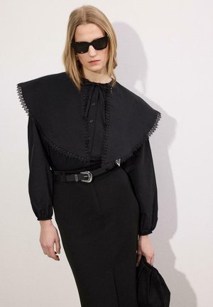 Blondynka w koszuli z dużym kołnierzem wykończonym czarną koronką, czarną ołówkową spódnicą, paskiem, okularami przeciwsłonecznymi, trzymająca dopasowaną czarną torebkę, stojąca na tle białej ściany.