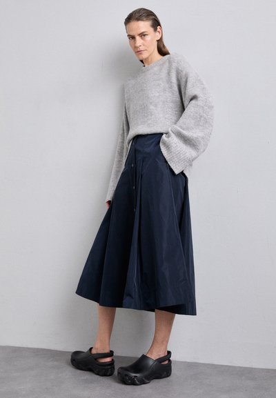 Pull en tricot gris, jupe midi marine avec des détails de boutons, et sabots noirs à semelles texturées. Silhouette simple et ample.