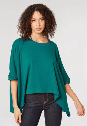 Tee-farbene, locker sitzende Bluse mit rundem Ausschnitt und kurzen Ärmeln. Der Stoff wirkt weich mit einem leicht schimmernden Finish. Der Saum ist asymmetrisch und fällt an den Seiten länger.
