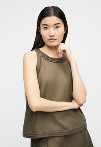 Νεαρή γυναίκα με μακριά σκούρα μαλλιά που φοράει ελιά αμάνικο τοπ με πλεκτό τελείωμα, πόζα με το ένα χέρι στο πιγούνι και το άλλο σταυρωμένο.