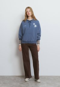 Sudadera con capucha azul con bolsillo frontal, gráficos de estrellas blancas y cordón ajustable. Combinada con pantalones marrones con botones laterales y una camiseta de rayas debajo.