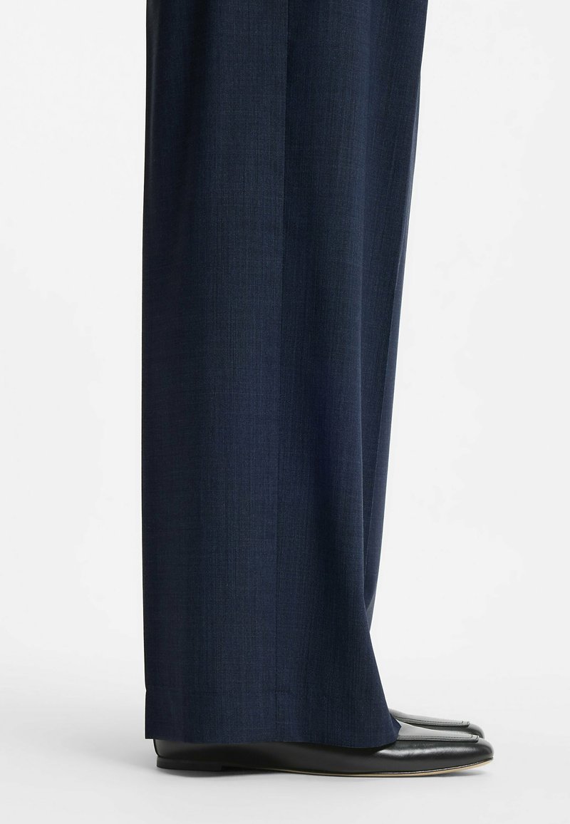 Donkere marineblauwe wijde trousers die zwarte schoenen met een vierkante neus bedekken op een lichte achtergrond, met zichtbare stoftextuur en pasvorm van het kledingstuk.