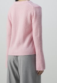 Ljusrosa, fluffig tröja med långa ärmar, rundad halsringning och ribbad kant. Materialet ser mjukt och något texturerat ut. Kombinerad med grå byxor.