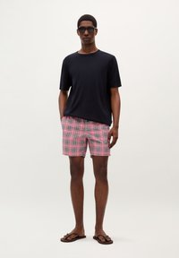 Tricou bleumarin cu guler rotund, pantaloni scurți de baie cu carouri roz și albe, ochelari de soare negri, sandale cu talpă subțire maro, stând pe un fundal simplu.