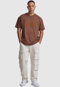Camiseta de algodón marrón con logotipo en relieve, combinada con pantalones cargo blancos que tienen múltiples bolsillos y puños ajustados, usados con zapatillas blancas.