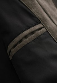 Gros plan sur un tissu noir avec une bordure en daim marron foncé et une fente de poche, montrant la texture et les détails de couture.