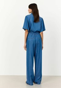 Denim kombinezon s opuštenim krojem, kratkim rukavima, elastičnom strukom i strukturiranim ramenima u srednje plavoj boji s suptilnim detaljima šivanja.