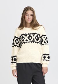 Maglione color crema con motivi geometrici neri e tessuto texturizzato. Caratterizzato da maniche lunghe e un colletto rotondo. Abbinato a pantaloni scuri.