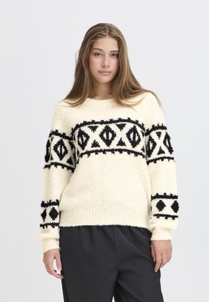 Maglione color crema con motivi geometrici neri e tessuto texturizzato. Caratterizzato da maniche lunghe e un colletto rotondo. Abbinato a pantaloni scuri.