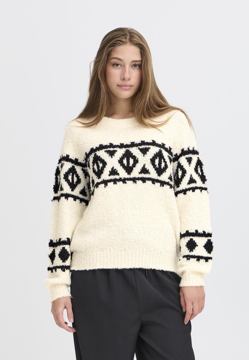 Maglione color crema con motivi geometrici neri e tessuto texturizzato. Caratterizzato da maniche lunghe e un colletto rotondo. Abbinato a pantaloni scuri.