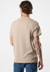 Beige T-Shirt mit kurzen Ärmeln aus gerippter Textur, mit Rundhalsausschnitt und gesteppten Schulternähten. Sichtbare Tattoos am Arm.