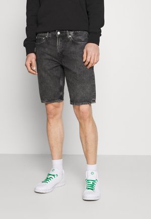 Mężczyzna ubrany w czarne, sprane szorty jeansowe, białe trampki z zielonymi sznurówkami, białe skarpetki oraz czarną bluzkę z długim rękawem, stojący na jednolitym tle.