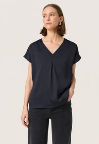 Blusa de manga corta azul marino con escote en V, tejido texturizado, caída suave y detalle de pliegue en la parte delantera. Usada con jeans negros.