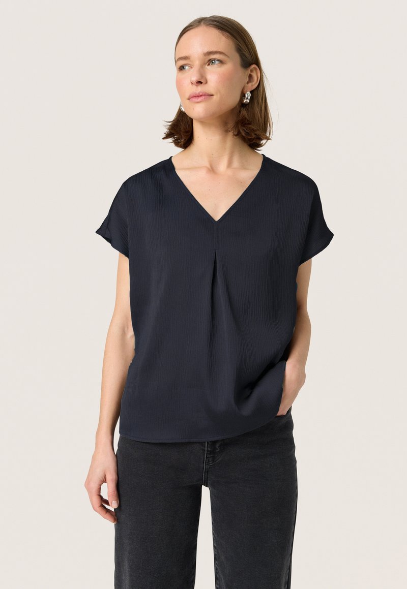 Blusa de manga corta azul marino con escote en V, tejido texturizado, caída suave y detalle de pliegue en la parte delantera. Usada con jeans negros.