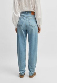 Osoba ubrana w jasnoniebieskie spodnie z wysokim stanem, białą, fakturowaną bluzkę z długim rękawem i brązowe buty, stojąca na jednolitym jasnym tle.