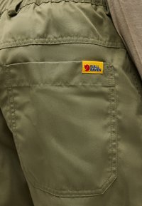 Κοντινή λήψη της πίσω τσέπης χακί παντελονιού με κίτρινη ετικέτα λογότυπου Fjällräven στην πάνω δεξιά γωνία της τσέπης.