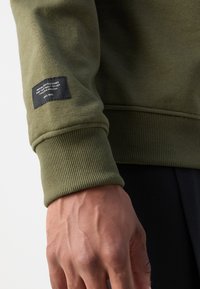 Olivengrønn genser med ribbestrikkede mansjetter og kant, som har en svart etikett på ermet med hvit tekst som beskriver merket og pleieinstruksjoner.