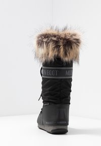 Botas de invierno altas negras con puño de piel marrón, marca "Moon Boot", etiqueta de impermeabilidad y suela en cuña sobre fondo blanco.