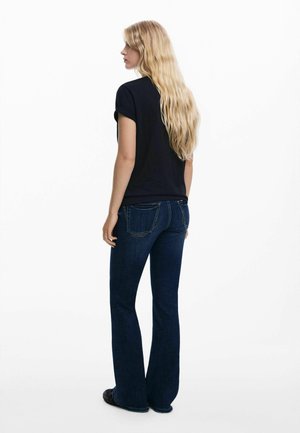 Black t-shirt with rolled sleeves, paired with flared dark blue jeans. Long wavy hair, viewed from the back against a white background.