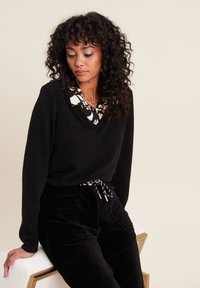 Schwarzer V-Ausschnitt-Pullover mit einer weichen Textur, über einem weißen Kragen mit einem schwarzen Blumenmuster getragen. Kombiniert mit schwarzen Cord-Hosen.