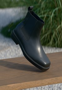 Melna ādas potīšu zābaku ar gludu virsmu, elastīgām malām un cilpiņas vilkšanas cilpu, kas atpūšas uz koka virsmas pret izplūdušu fonu.