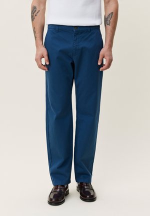 Mann iført blå bukser med rette ben, hvit skjorte, mørkebrune loafers, med synlige tatoveringer på begge underarmer.