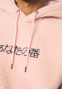 Lichtroze fleece hoodie met een gestructureerd oppervlak, trekkoorden met metalen uiteinden en zwarte geborduurde tekst aan de voorkant.