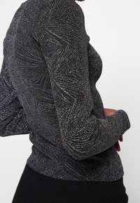 Nahaufnahme einer Person, die ein schwarzes Langarmshirt mit silbernen glitzernden Wirbelmustern und schwarze strukturierte Hosen vor einem einfarbigen Hintergrund trägt.