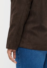 Giacca in finta suede marrone con una texture morbida, maniche aderenti e una silhouette pulita. Indossata con jeans di denim blu, con la mano appoggiata sul fianco.