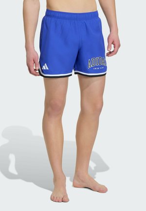 Man draagt blauwe Adidas zwemshorts met witte en zwarte accenten en logo's, staand op blote voeten tegen een lichte achtergrond.